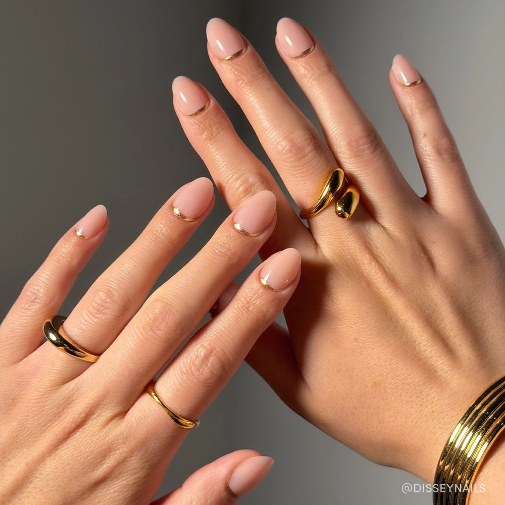Close-up of hands wearing glamnetic Glam Goals short oval press-on nails in nude pink with gold crescent accents, paired with chunky gold rings and a bracelet for a chic finish. - Product image