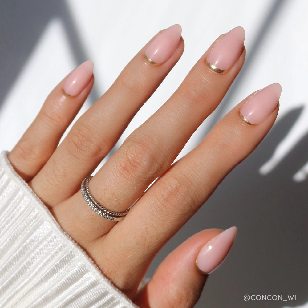 A hand with short almond nails in glamnetic’s Goal Chaser, a light pink polish with minimalist gold half-moon and French tip designs. Two thin, stacked rings with small gems are worn. The white ribbed sleeve completes the softly lit look. - Product image