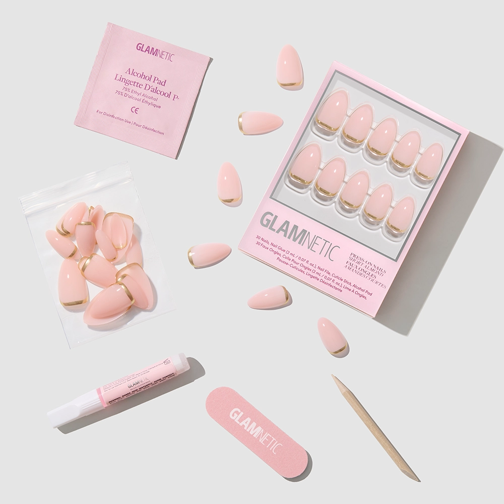 Flat lay of the glamnetic Goal Chaser pink press-on nail kit, with short almond and gold French tip nails, plus a nail file, cuticle stick, glue, alcohol pad, and extra nails on a light gray background. - Product image