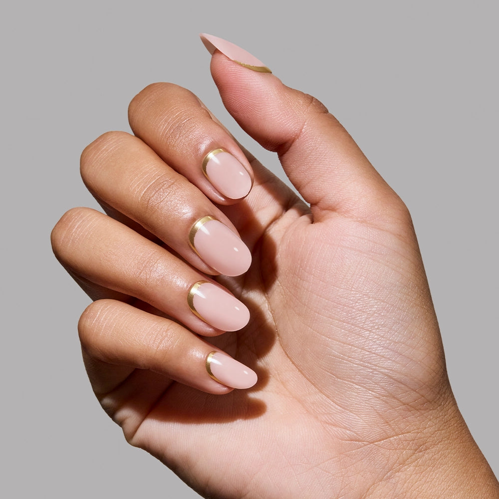 A hand with almond-shaped nails painted in glamnetic's "Goal Digger," a nude pink shade with thin metallic gold accents, reminiscent of gold foil nails, shown against a plain gray background. - Product image