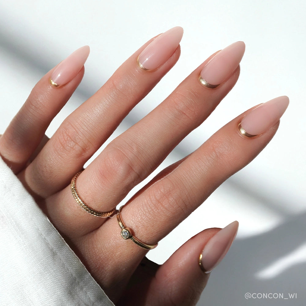 A hand with medium almond nails in glamnetic's Goal Getter—light nude polish with thin gold French tips—wears two gold rings against a softly lit white background. - Product image