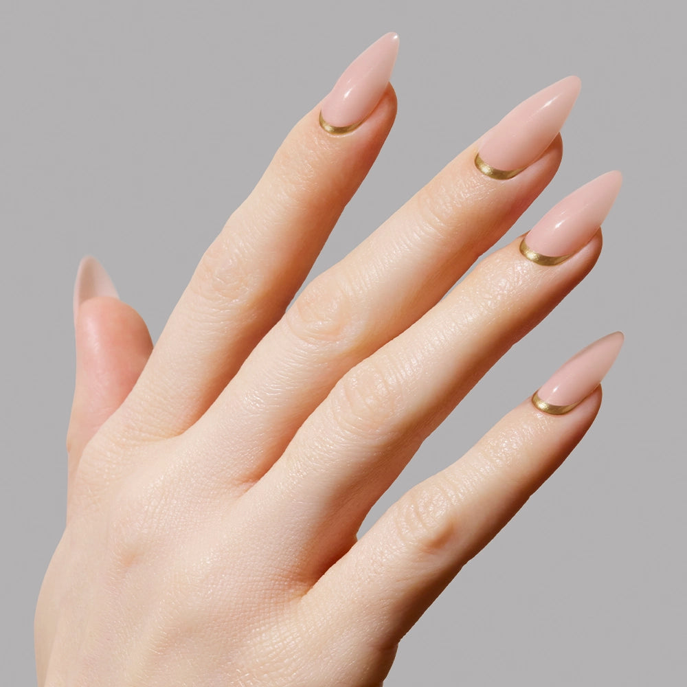 A hand wears glamnetic’s Goal Getter medium almond nude press-on nails, each with a minimalist gold accent near the cuticle, displayed against a plain light gray background. - Product image