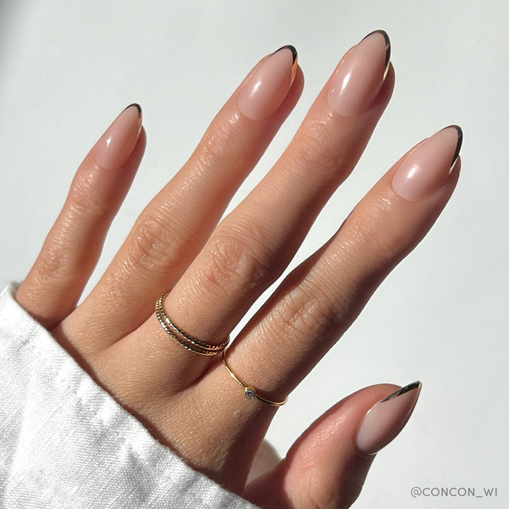 A hand with almond-shaped Glamnetic Gold Standard nails—nude base, thin black French tips—displays three gold rings and a white long-sleeve top against a softly lit plain background.