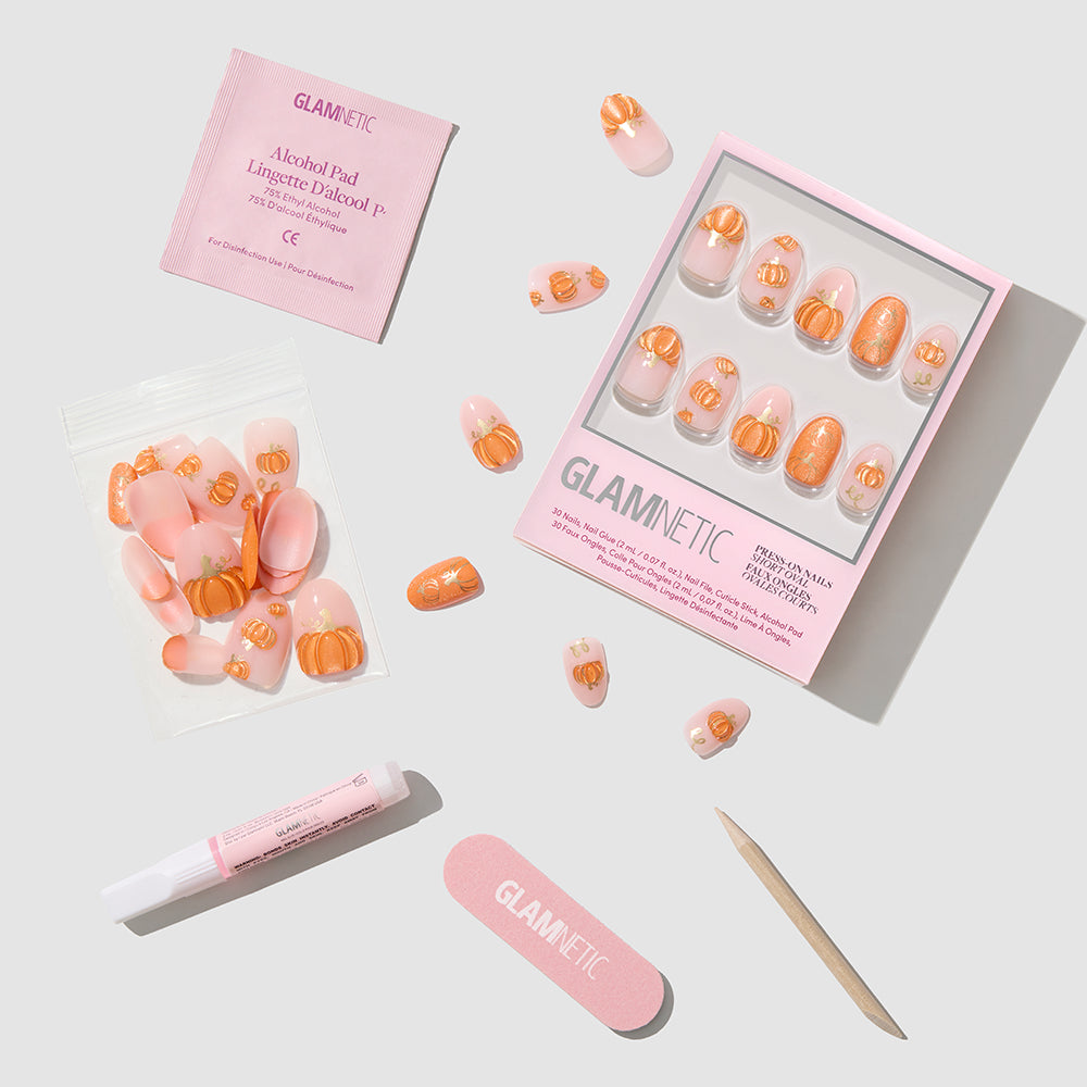 A glamnetic Golden Gourds press-on nail kit featuring short oval nails adorned with pink and orange pumpkin nail art, including glue, a nail file, a cuticle stick, an alcohol pad, and extra nails—arranged neatly on a white background. - Product image