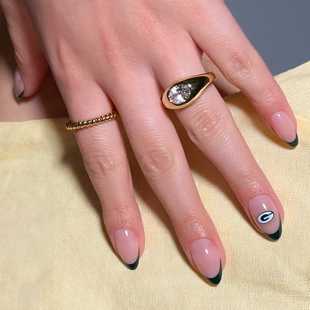 A hand wearing glamnetic’s NFL Press-On Nails—styled in an almond shape with a French tip design and a Green Bay Packers logo on the ring finger—adorned with two gold rings, rests on yellow fabric. - Product image
