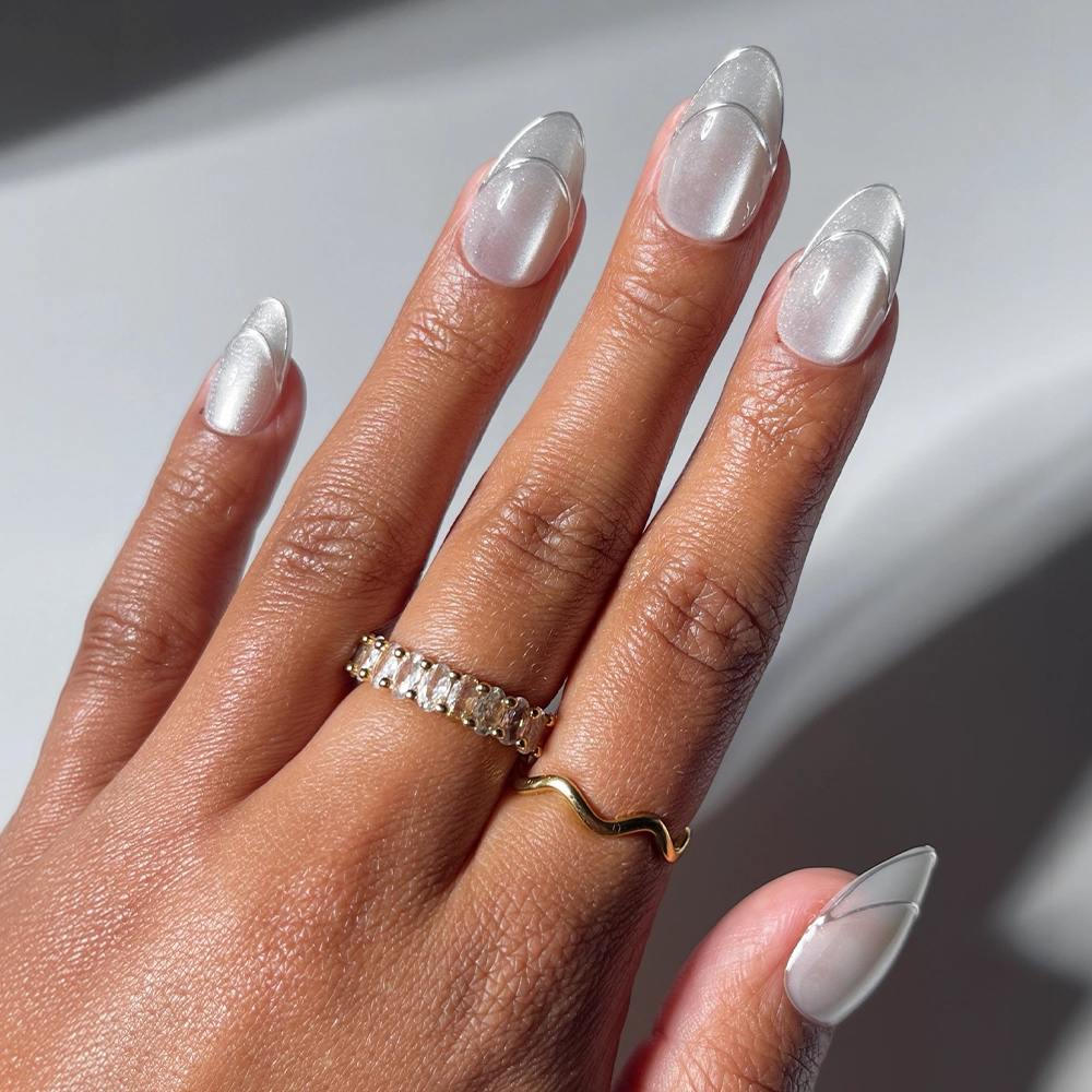 A hand with short almond nails, painted in “Heavenly Metal” by glamnetic—a sheer milky white press-on set outlined in silver shimmer—is adorned with two gold rings, one wavy and the other set with rectangular gemstones. The softly lit, neutral background complements the elegant glamnetic nails beautifully. - Product image
