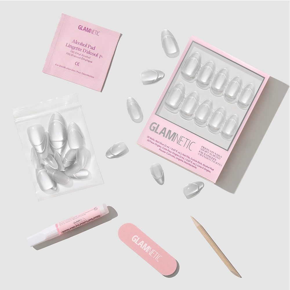 Introducing glamnetic’s Heavenly Metal press-on nail kit: a stylish set of clear press-on nails packaged in a signature pink box, complete with nail glue, alcohol pad, wooden stick, file, and extra nails. Perfect for creating your favorite short almond look or adding silver shimmer accents—all beautifully arranged on a light gray background. - Product image