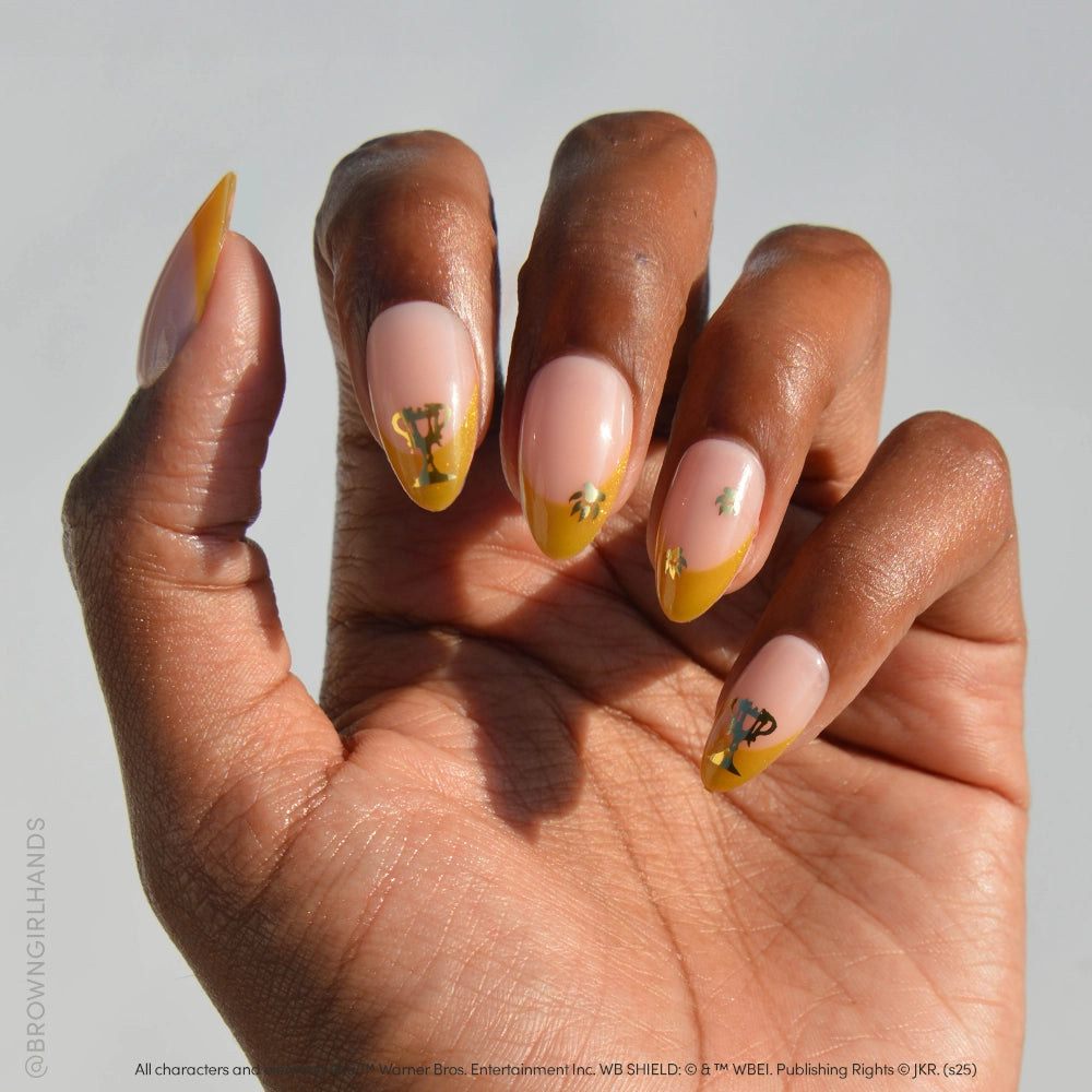 A hand with medium brown skin displays long, almond nails in a nude base and yellow velvet French tips, each decorated with gold and green Helga Hufflepuff™ designs from glamnetic.