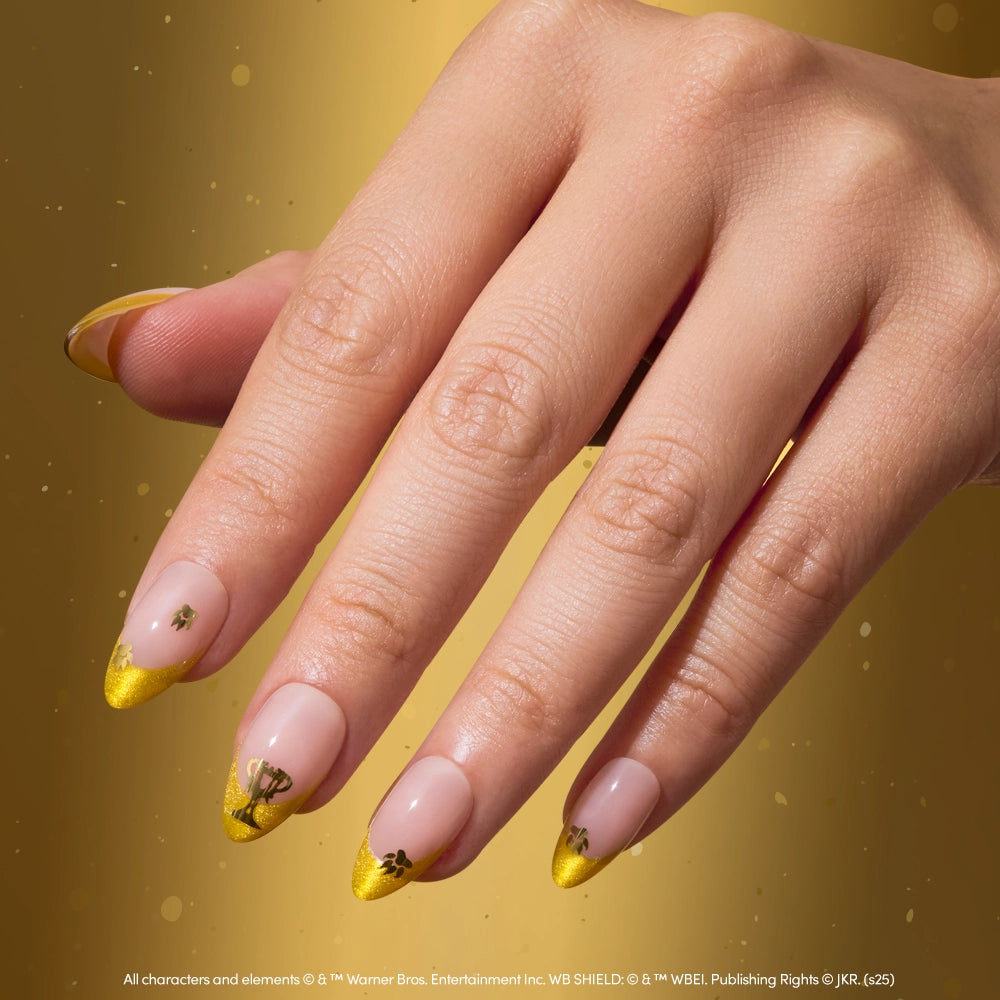 A hand with long, almond-shaped Helga Hufflepuff™ nails by glamnetic features a nude base and yellow velvet French tips with small black designs. The background shimmers in a matching gold hue. - Product image