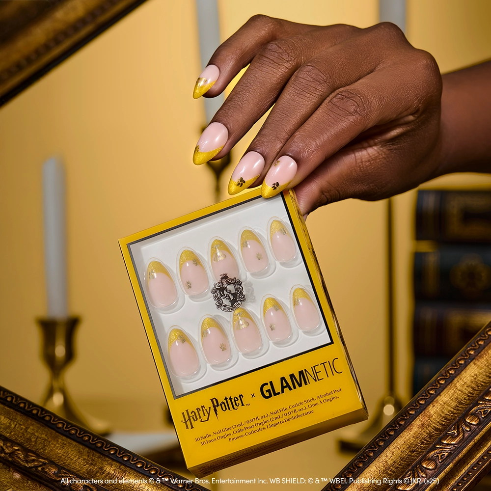 A hand with long, almond-shaped nails in yellow velvet French tips holds a box of glamnetic’s Helga Hufflepuff™ set. In the background are a gold frame, candles, books, and a magical creatures sticker sheet for whimsy. - Product image