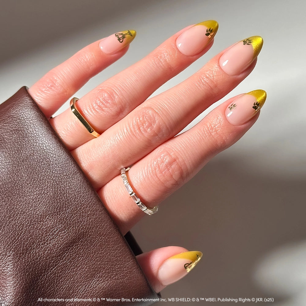 A hand with manicured nails featuring a nude base, yellow velvet French tips inspired by the glamnetic Helga Hufflepuff™, and small black designs, adorned with two rings, rests on a brown leather surface. - Product image