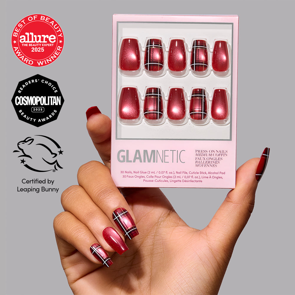 A hand holds a box of glamnetic Holiday Tartan press-on nails, featuring a vibrant red and black plaid design, with matching medium coffin nails applied. The Allure, Cosmopolitan, and Leaping Bunny certification logos are displayed on a gray background. - Product image