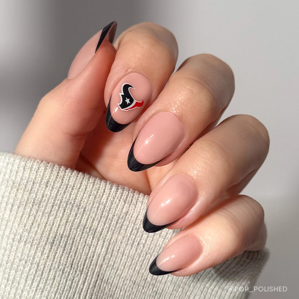 A hand shows the Houston Texans Press-On Nail Set by glamnetic, with an almond manicure: nude base, black French tips, and one nail featuring a detailed Houston Texans logo. The person wears a light gray textured sleeve.