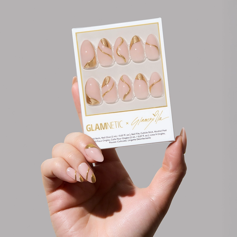 A hand holds up the Glamnetic x Glamzilla nail kit, revealing 30 press-on nails in varying abstract gold designs on a nude-pink base. The box features Glamnetic branding and Glamzilla’s signature.