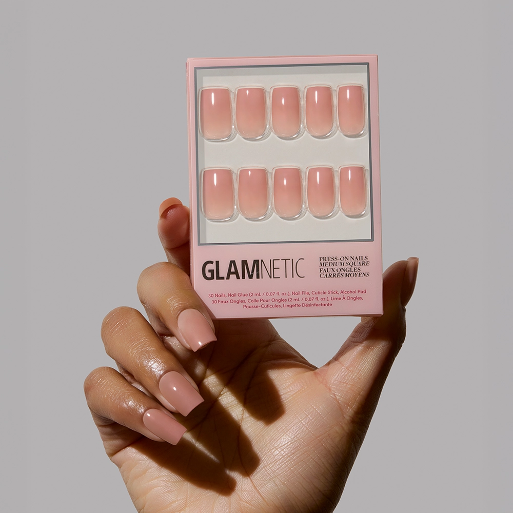 A hand with long, glossy, nude-pink ombré nails holds a box of glamnetic In The Nude press-on nails against a plain light gray background, highlighting the perfect minimalist manicure. - Product image