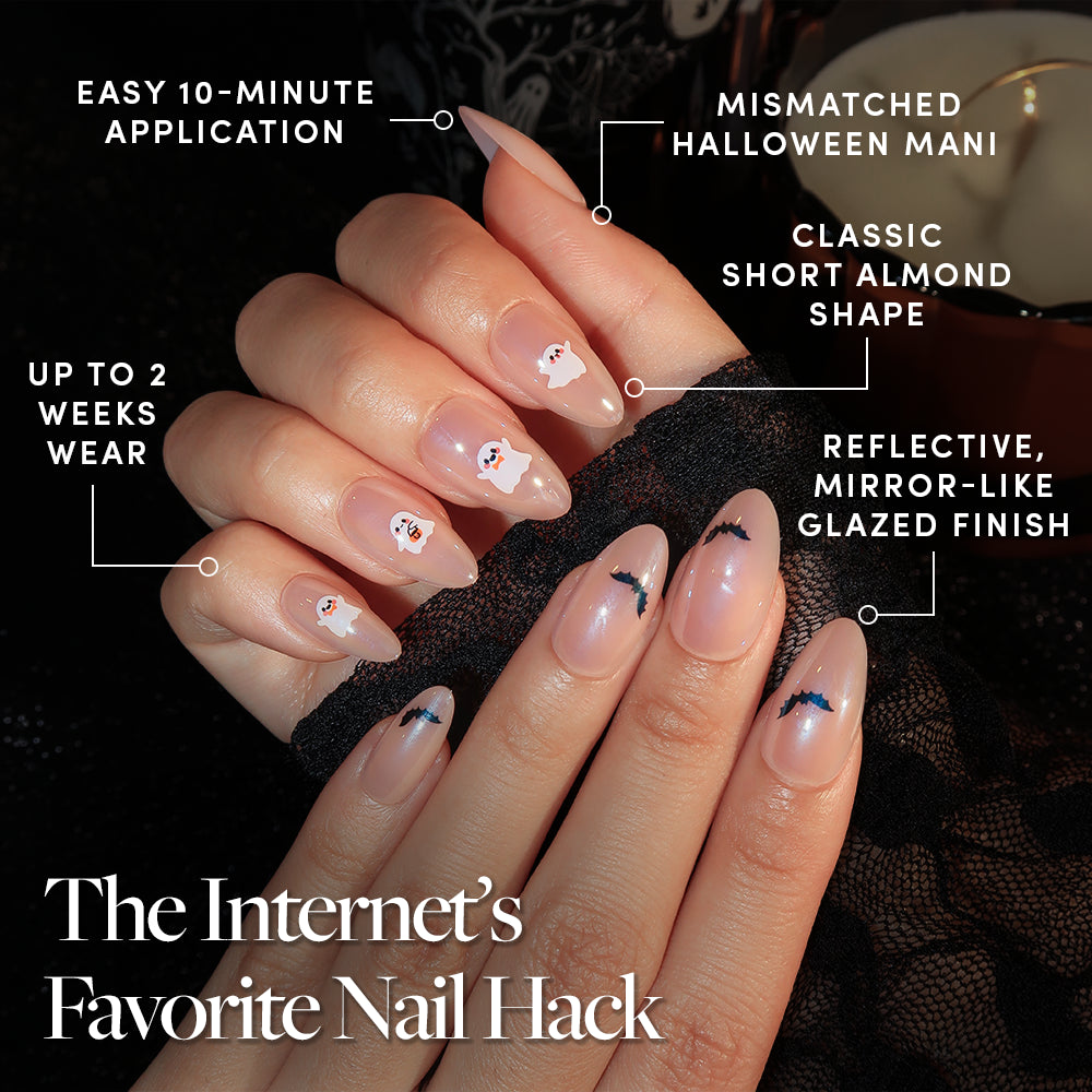A hand with short almond-shaped glamnetic In The Spirit press-on nails, featuring a mismatched Halloween ghost and bat design, is shown. Text highlights benefits: 10-minute application, lasts 2 weeks, classic shape, and a glossy glazed finish. - Product image