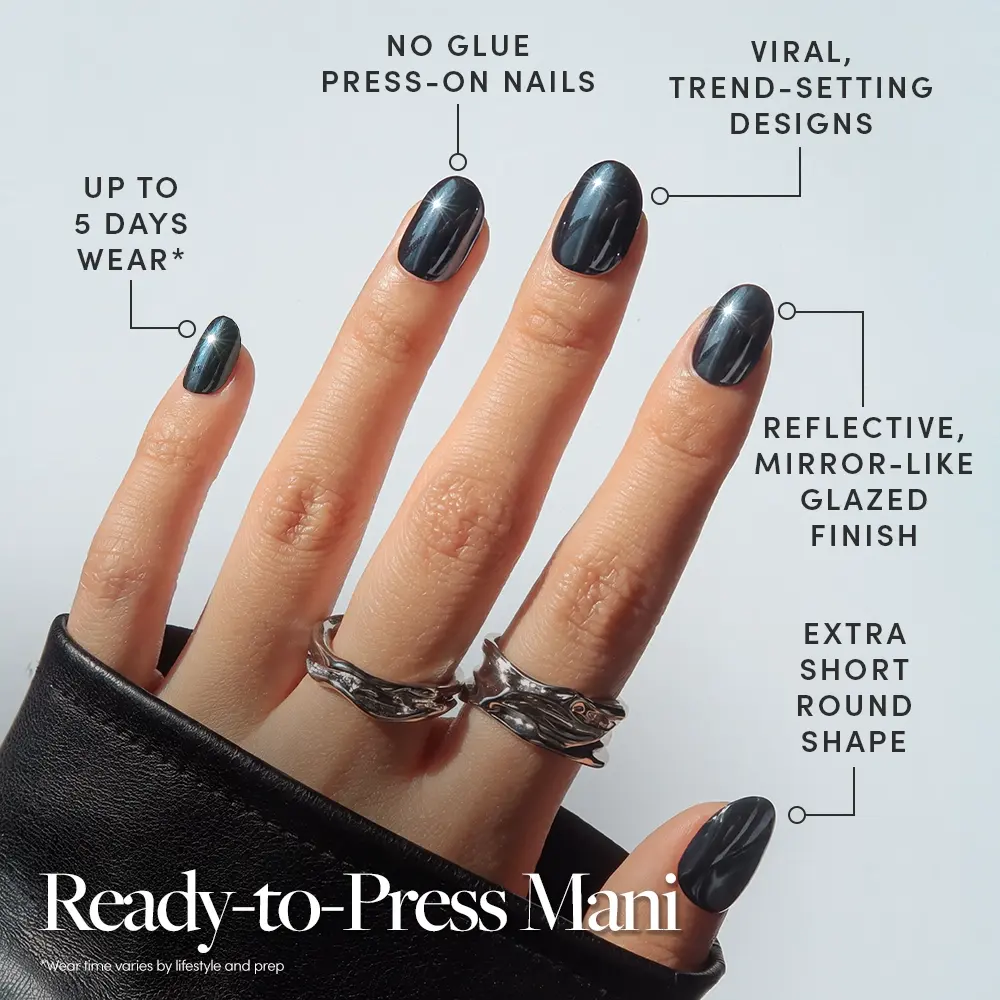 A hand showcases glamnetic’s Ink Spell press-on nails in a shiny, dark finish, with labels pointing out key features: no glue needed, up to 5 days of wear, reflective glaze, and an extra short round shape. The hand is adorned with silver rings and a black sleeve. Text: Ready-to-Press Mani—experience the effortless beauty of stick-on nails. - Product image
