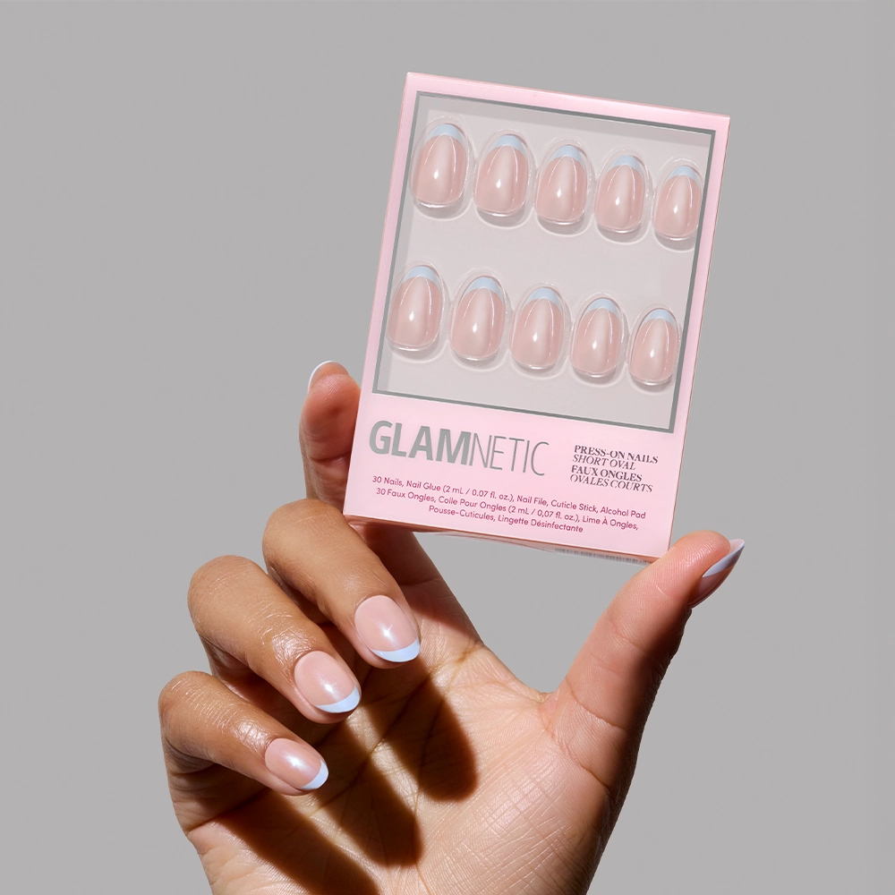 A hand with a natural pink and light blue French manicure holds a glamnetic Into Blue press-on nail set box against a plain gray background. The salon-quality packaging showcases ten nails with periwinkle micro French tips inside. - Product image