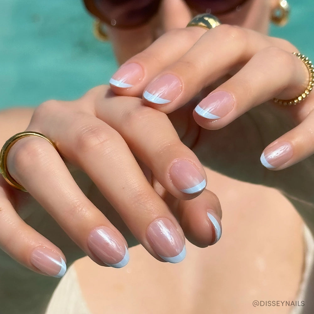 A close-up of hands wearing glamnetic’s Into Blue: salon-quality periwinkle micro French tips on short, natural nails, paired with gold rings and sunglasses against a blurred turquoise background. - Product image