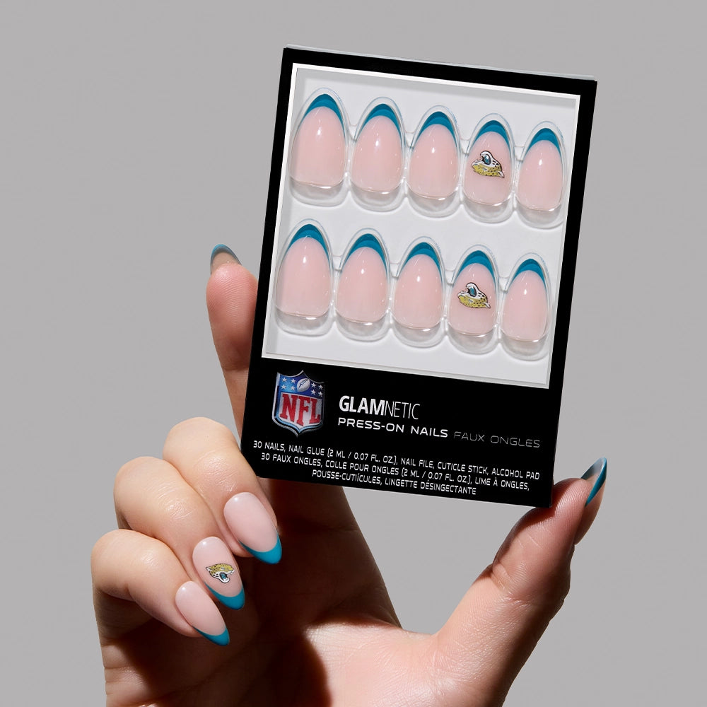A hand with blue French tip nails holds the glamnetic Jacksonville Jaguars Press-On Nail Set, featuring the team's logo on accent nails. The packaging shows ten blue-tipped press-ons, perfect for a sporty almond manicure.