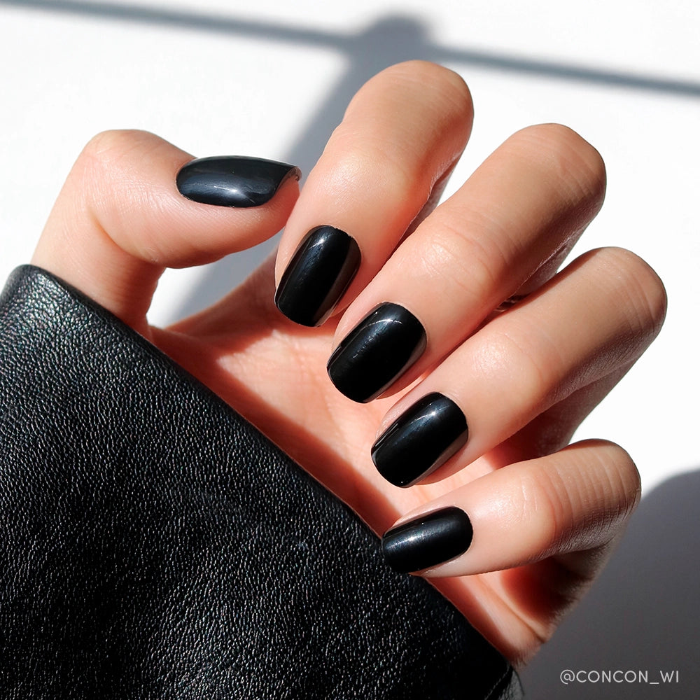 A hand with glossy squoval nails in glamnetics Jet Black, partially covered by a black leather sleeve, stands out against a bright white background with soft shadows. - Product image