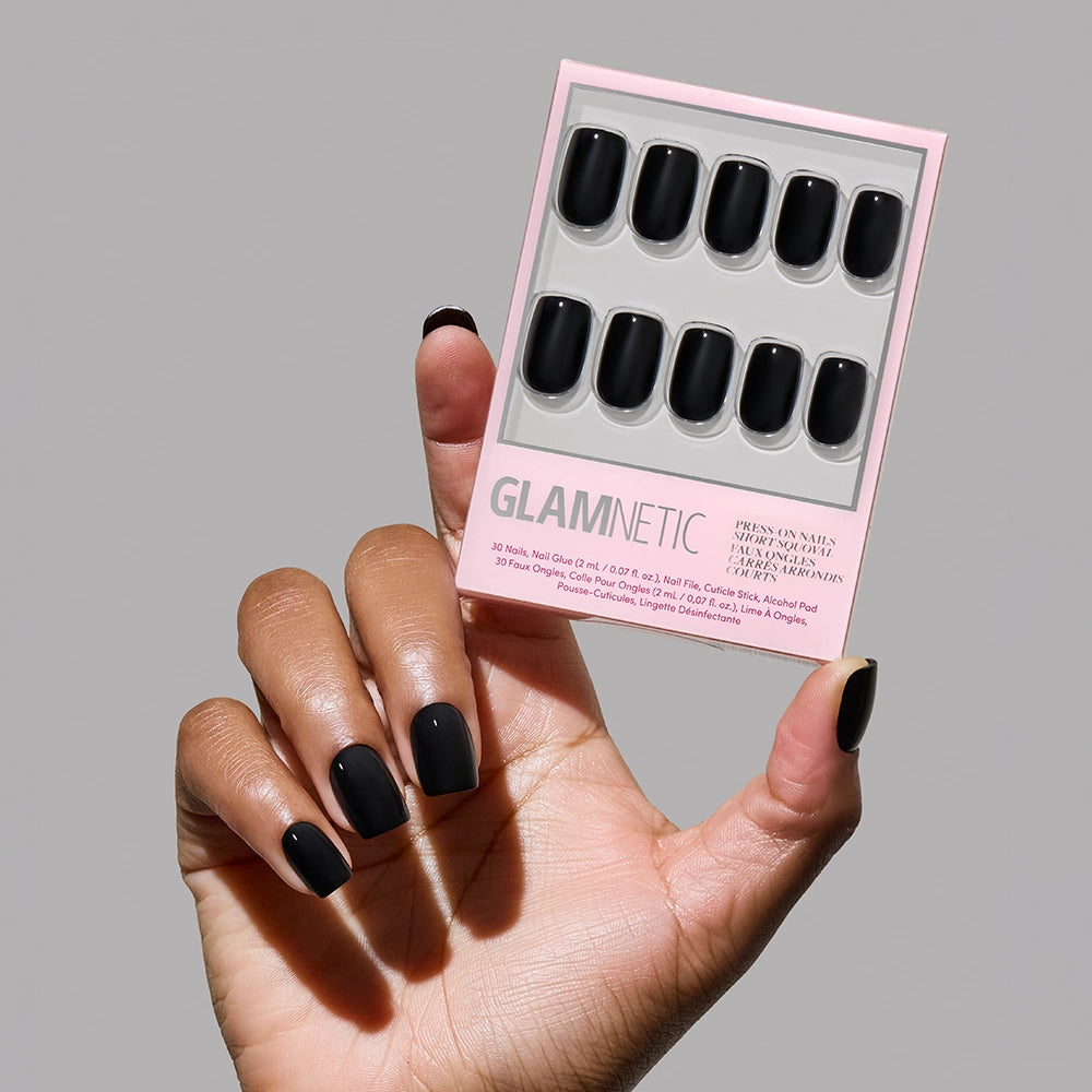 A hand with glossy squoval nails holds a glamnetic box, showcasing the Jet Black reusable vegan nail set—perfectly matching nails—against a plain gray background.
