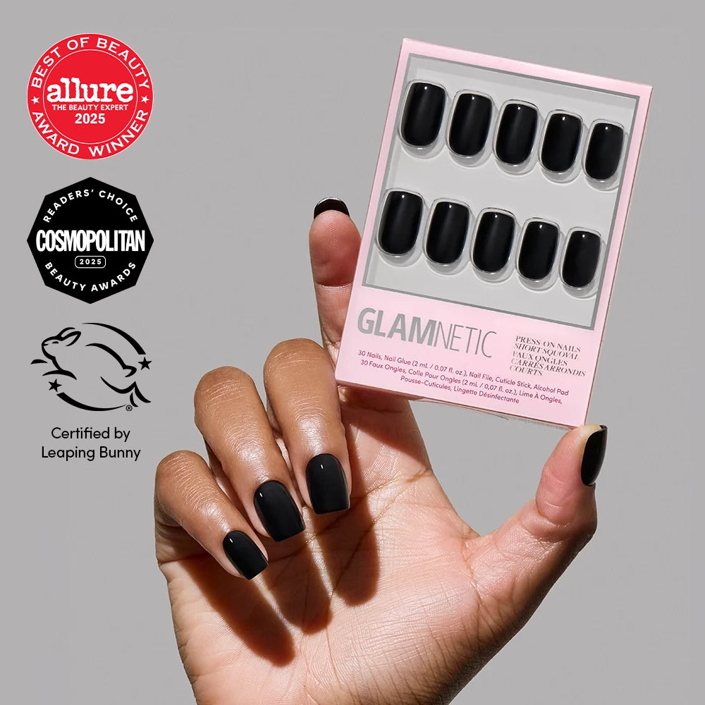 A hand with glossy Jet Black press-on nails from glamnetic holds a box of matching nails. Award badges from Allure, Cosmopolitan, and Leaping Bunny are shown on the left.