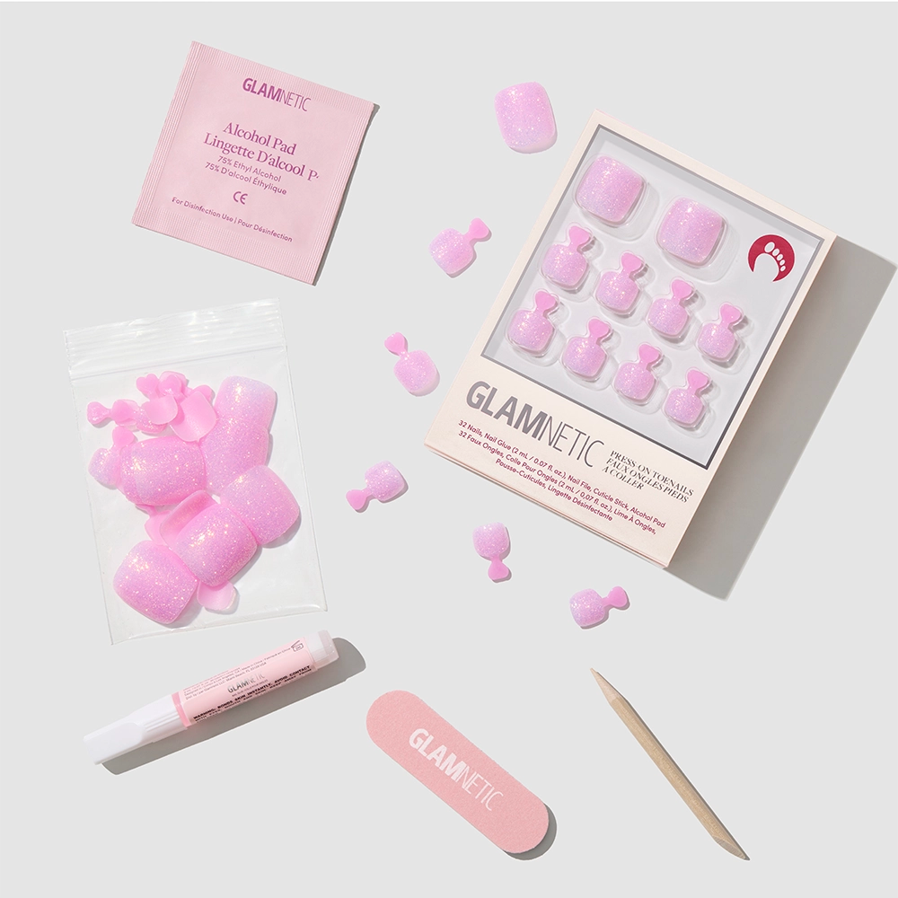 The glamnetic Juicy Toenail set features vegan, cruelty-free pink glitter press-on toenails with adhesive tabs, a nail file, cuticle stick, glue tube, and alcohol pad—all displayed neatly on a light gray background. - Product image