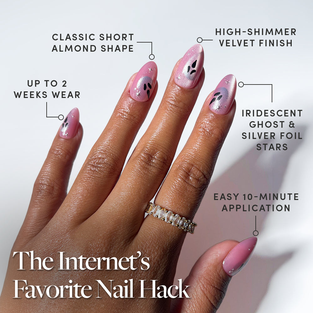 A hand showcases “Just A Ghoul” by glamnetic—short almond, pink shimmery press-on nails adorned with black and silver star designs, iridescent foil accents, and a subtle velvet finish. Enjoy salon-quality nails at home with an easy 10-minute application and up to 2 weeks of flawless wear. - Product image