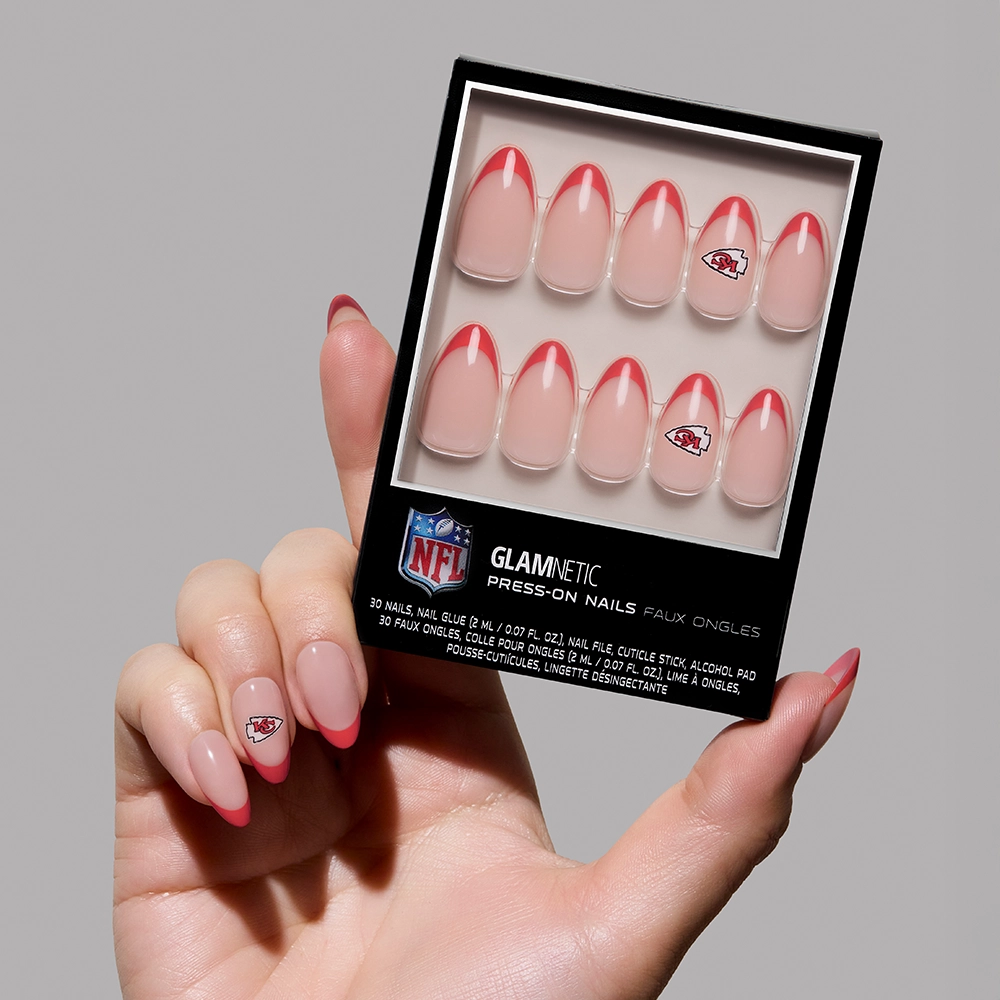 A hand holds the glamnetic Kansas City Chiefs Press-On Nail Set, featuring a red French tip design and NFL logo nails. The person is wearing one of the Chiefs press-ons.