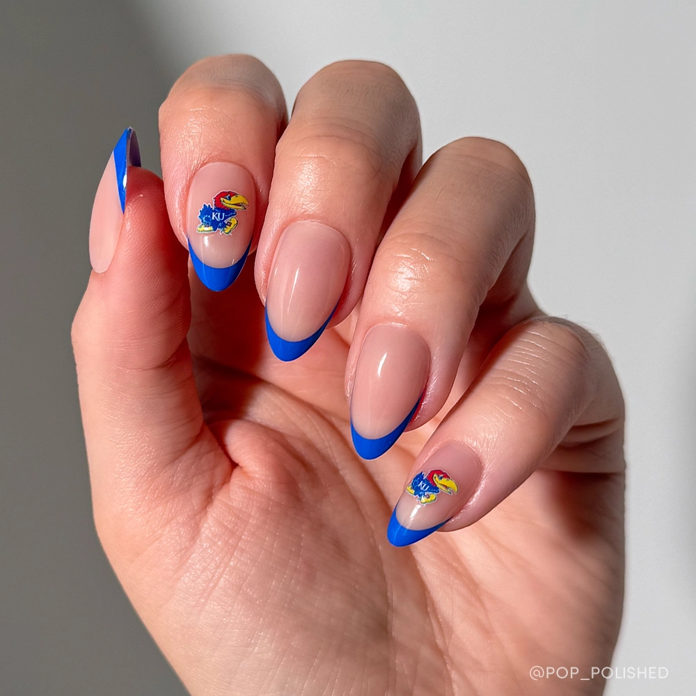 A hand with almond-shaped nails showcases a nude base, bright blue French tips, and two nails adorned with the glamnetic Kansas Jayhawks Press-On Nail Set. The well-lit image highlights the fan-inspired nail art.