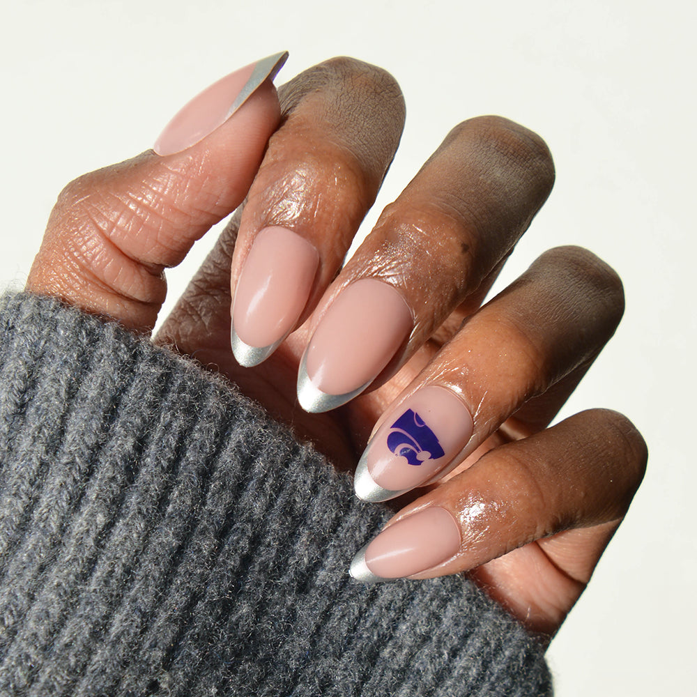 A hand is shown wearing glamnetic Collegiate Press-On Nails, featuring almond-shaped nails painted nude with silver tips and a purple wildcat logo accent on the ring fingernail. The person is dressed in a gray knit sweater. - Product image