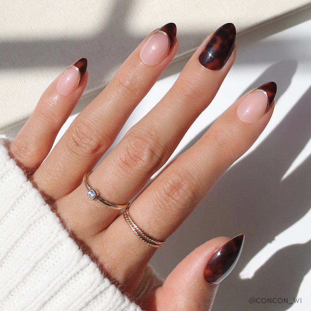 A hand with elegant almond-shaped nails showcasing alternating designs: glossy tortoiseshell full coverage and nude base with tortoiseshell French tips outlined in gold. The person is wearing minimalist gold rings and a cozy white sweater.