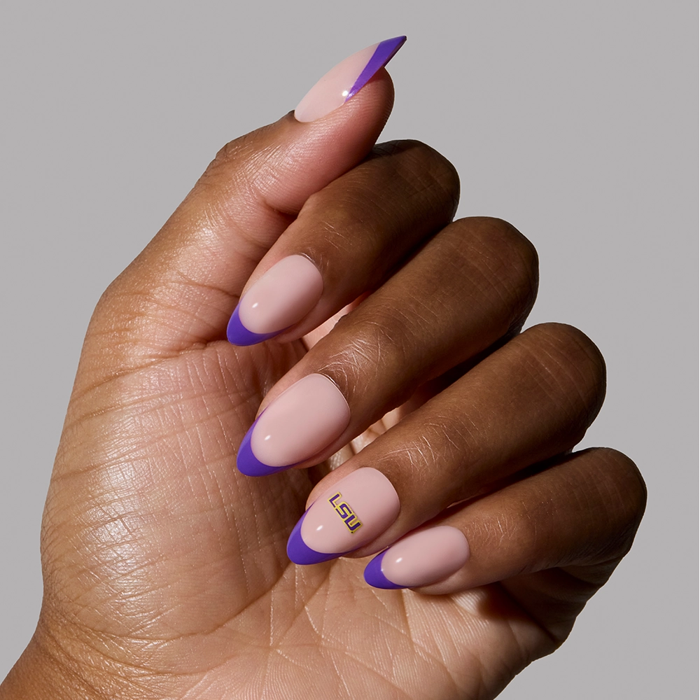 A hand with almond-shaped nude nails and purple French tips, plus one nail featuring gold LSU letters near the cuticle. Showcasing the officially licensed glamnetic LSU Tigers Press-On Nail Set. The background is light gray. - Product image