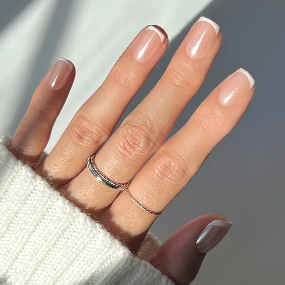 A hand with a light skin tone models glamnetic’s La Perle French tip nails in a chic short squoval shape, paired with two slim rings on the middle finger and a cozy white knit sweater.