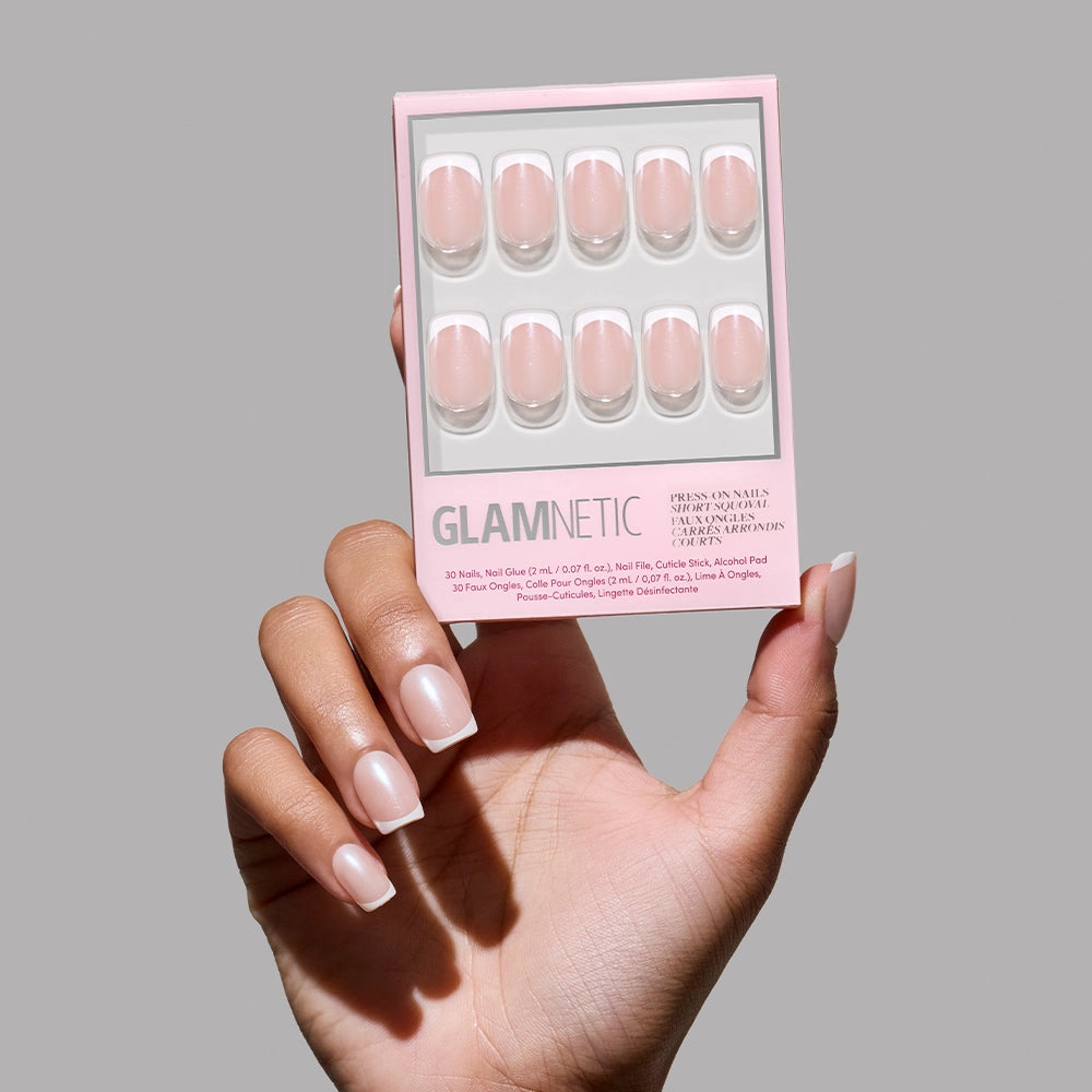 A hand with manicured nails holds a box of glamnetic La Perle press-on nails in light pink, featuring ten short squoval nails arranged in two rows against a plain gray background.