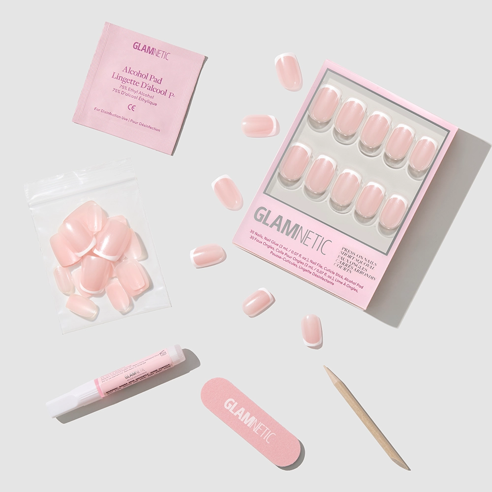 A flat lay showcases the glamnetic La Perle Nail Set with short squoval French tip nails, extra nails, a pink nail file, cuticle stick, glue tube, and alcohol pad arranged on a light gray background. - Product image