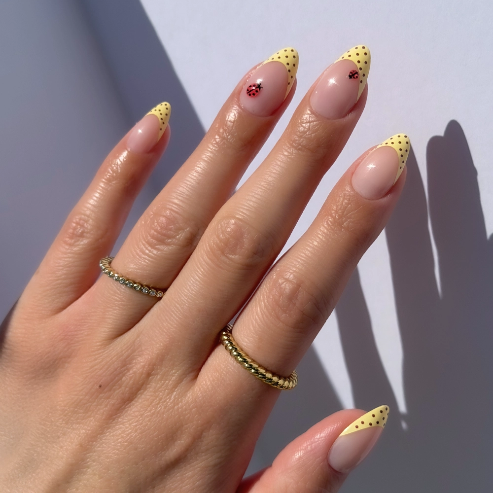 A hand with short almond nails showcases glamnetic's Lady Luck press-ons—yellow French tips with black polka dots and a small red flower on two nails. The person sports two gold rings, highlighting their trendy manicure.