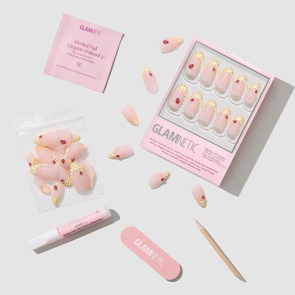 The glamnetic Lady Luck short almond press-on nails feature a pink and gold polka dot design and come with a wooden cuticle stick, nail file, glue, alcohol pad, and extra nails—ideal for an easy summer manicure on a light gray background. - Product image