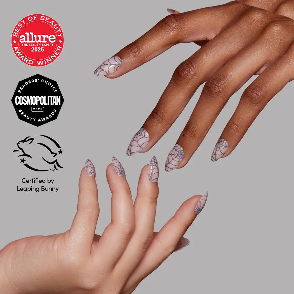 Two hands adorned with long, pointed, nude nails featuring the “Lady Web” white spiderweb nail art by glamnetic—perfect for Halloween nails—are showcased. Award badges from Allure, Cosmopolitan, and Leaping Bunny certifications appear on the left side of the image.