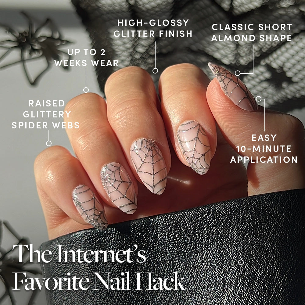 A hand displays Lady Web by glamnetic—almond-shaped press-on nails with a high-gloss, glittery spider web design—ideal for black spiderweb nails or a striking Halloween manicure. The text highlights their 2-week wear, 10-minute application, and other key features. The bold almond mani is shown resting on a black leather surface. - Product image