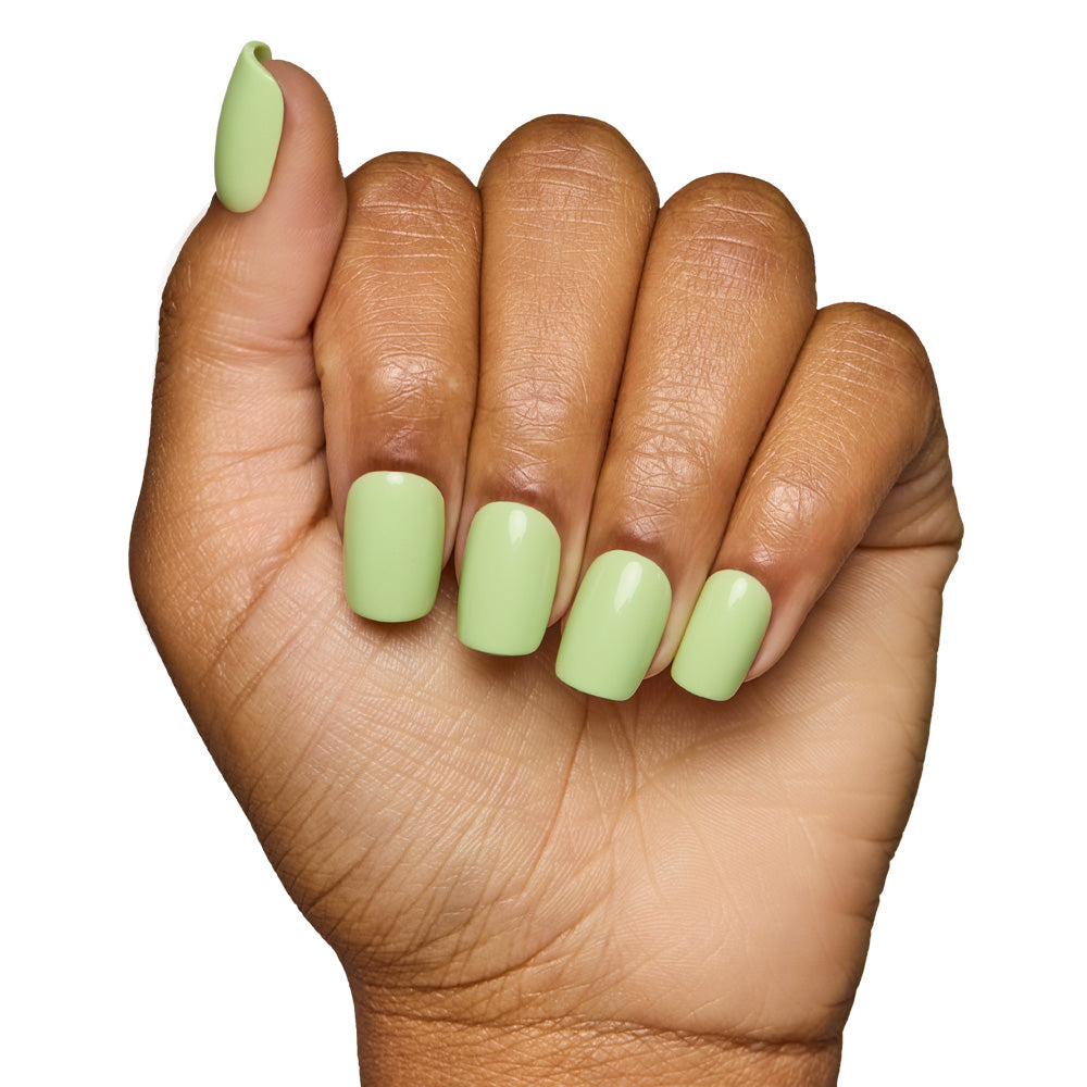 A hand with medium brown skin shows fingers curled inward, featuring neatly manicured short squoval nails painted in glamnetics Light Sage, a glossy pale green shade. The background is plain white. - Product image