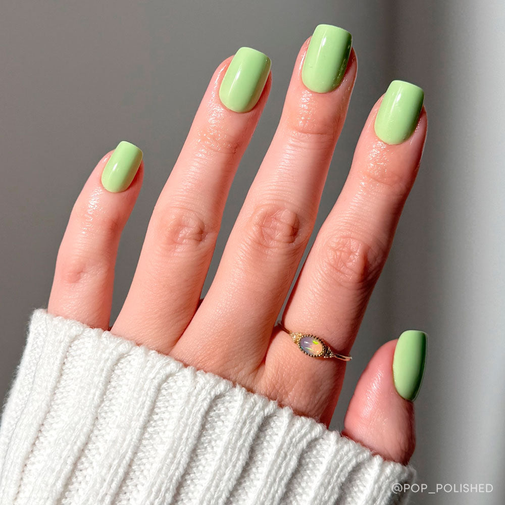 A hand with short squoval nails painted in glamnetic’s Light Sage shade, featuring a cream sweater and a delicate gold ring with a small oval gemstone. - Product image