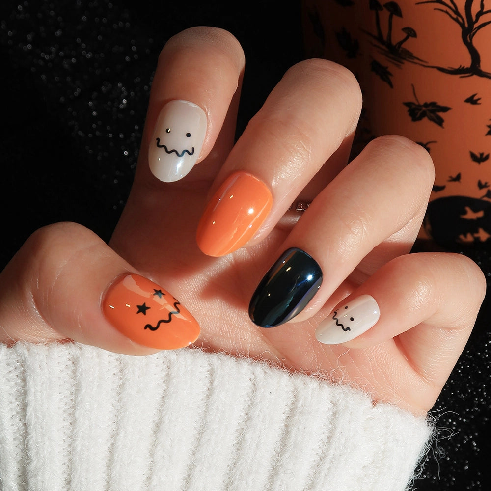 Close-up of a hand wearing glamnetic Lil Gremlins Halloween-themed press-on nails in a short oval shape. The set features solid orange and black nails with a glazed finish, as well as cream-colored nails adorned with cute, hand-drawn ghost and pumpkin faces in black ink. The person is dressed in a white sweater. - Product image