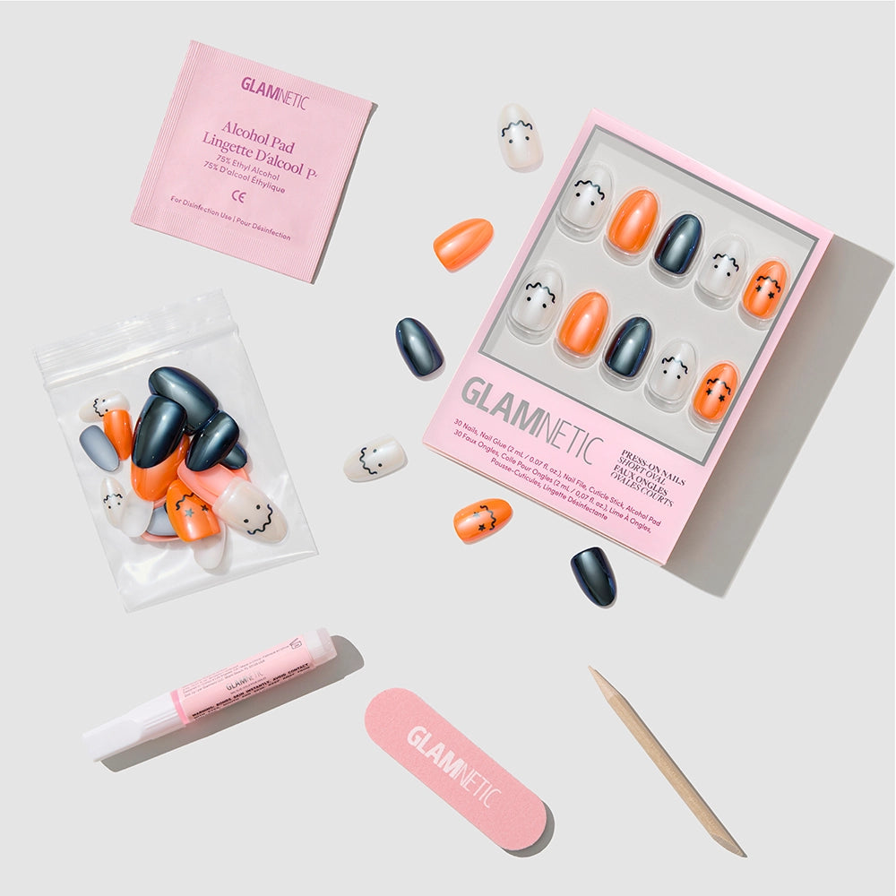 Flat lay of glamnetic Lil Gremlins short oval press-on nails featuring Halloween-themed ghost designs in orange, black, and white with a glazed finish; includes nail glue, wooden cuticle stick, nail file, alcohol pad, and extra nails displayed on a white background. - Product image