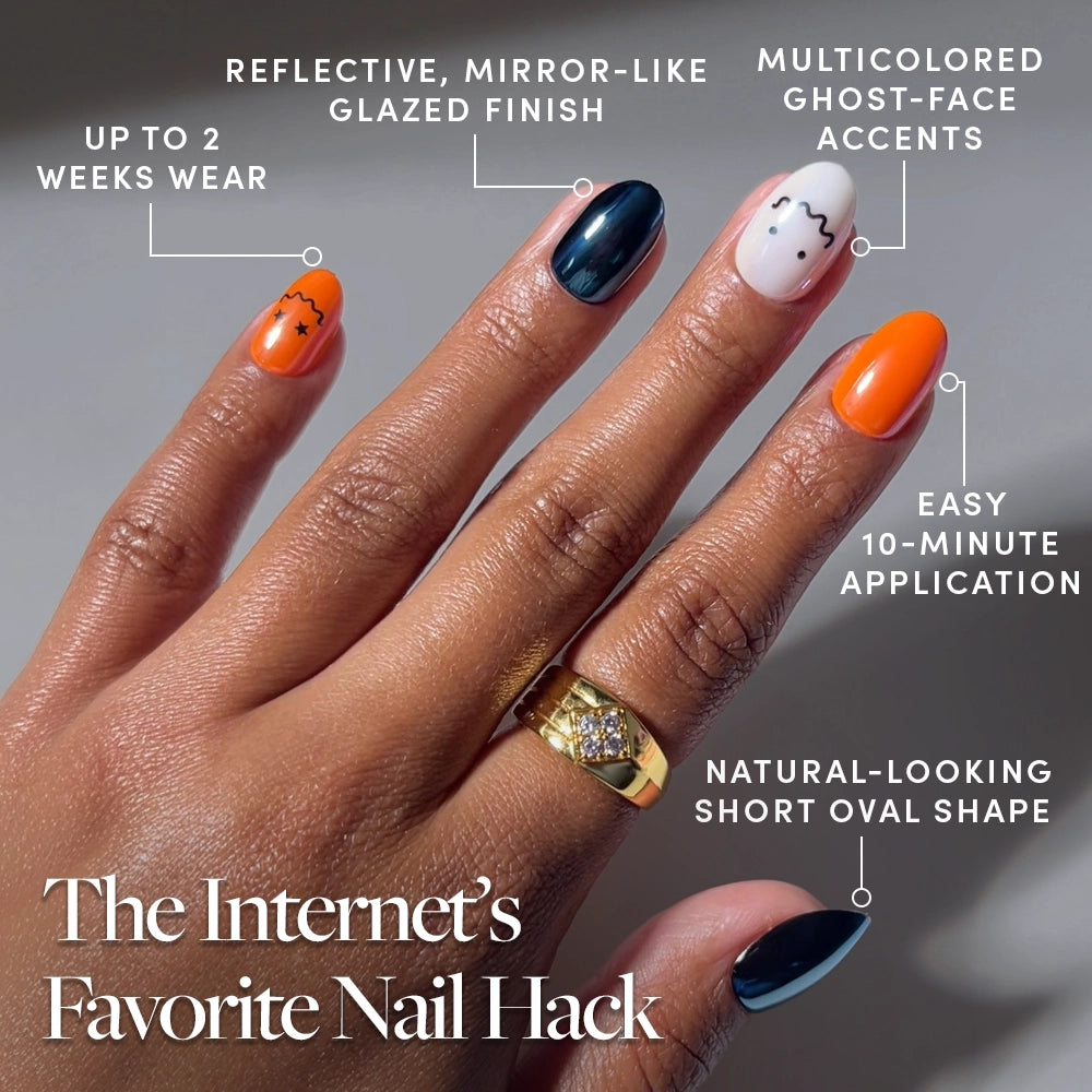 A hand showcases Lil Gremlins by glamnetic—multicolored, vibrant orange press-on nails in a natural short oval shape with ghost-face accents and a reflective glazed finish. Enjoy trendy nails with up to 2 weeks of wear and a quick 10-minute application. - Product image