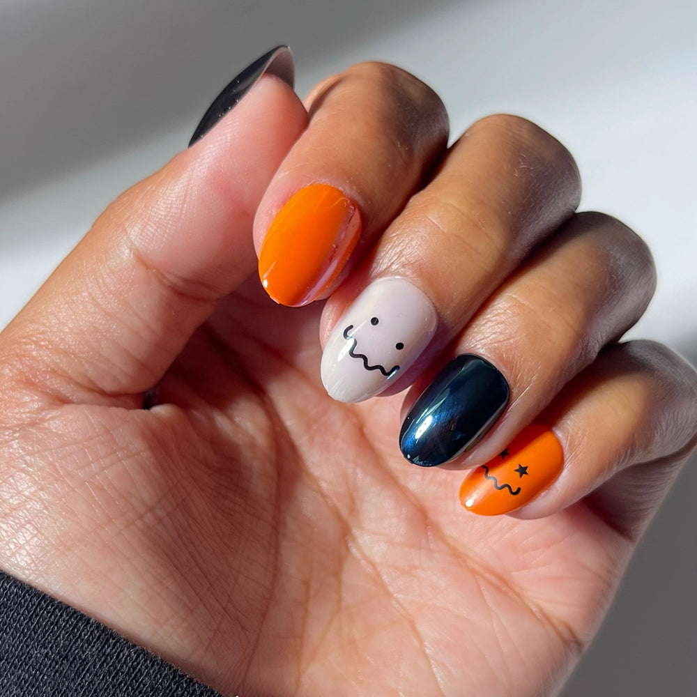 A hand shows off glamnetic’s Lil Gremlins press-on nails: short oval Halloween-themed nails in orange, black, and white, some adorned with ghost faces and a star. The set features a subtle glazed finish for extra shine. - Product image
