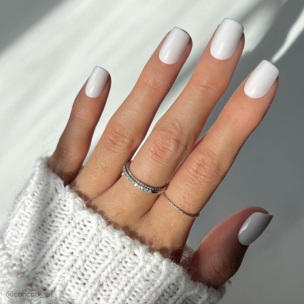 A hand in a white sweater sleeve displays glossy Lilac Gray squoval nails by glamnetic, complemented by two thin silver rings on the middle and ring fingers. The soft lighting adds elegance to the overall look. - Product image