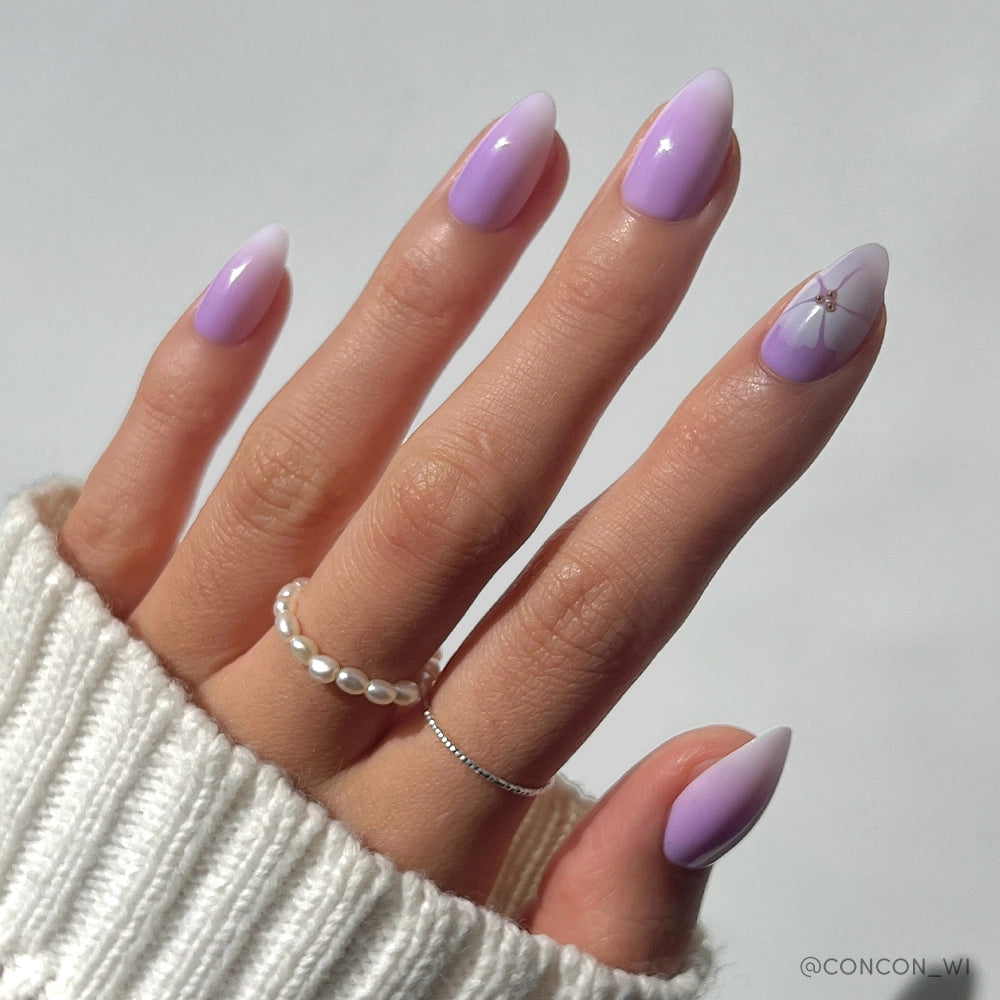 A hand with long almond nails in a lavender ombré, one featuring a delicate flower, styled with two rings, a white knit sweater, and the glamnetic Lilac Reef Press-On Nail Set for effortless style. - Product image