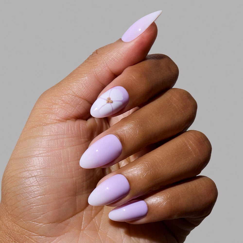 A hand with almond-shaped nails painted pastel lavender and white, featuring a floral design on the ring finger. Set against a plain gray background, this showcases the elegant Lilac Reef Press-On Nail Set by glamnetic. - Product image